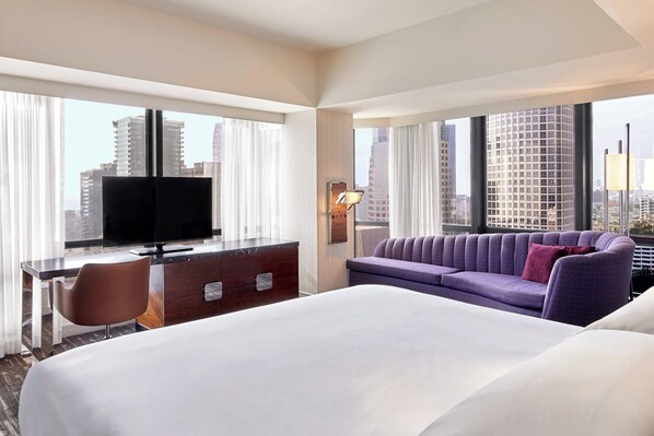 A hotel room with a large bed, a desk with a television, a purple sofa, and a city view through large windows. - Sheraton Grand Los Angeles (Los Angeles)