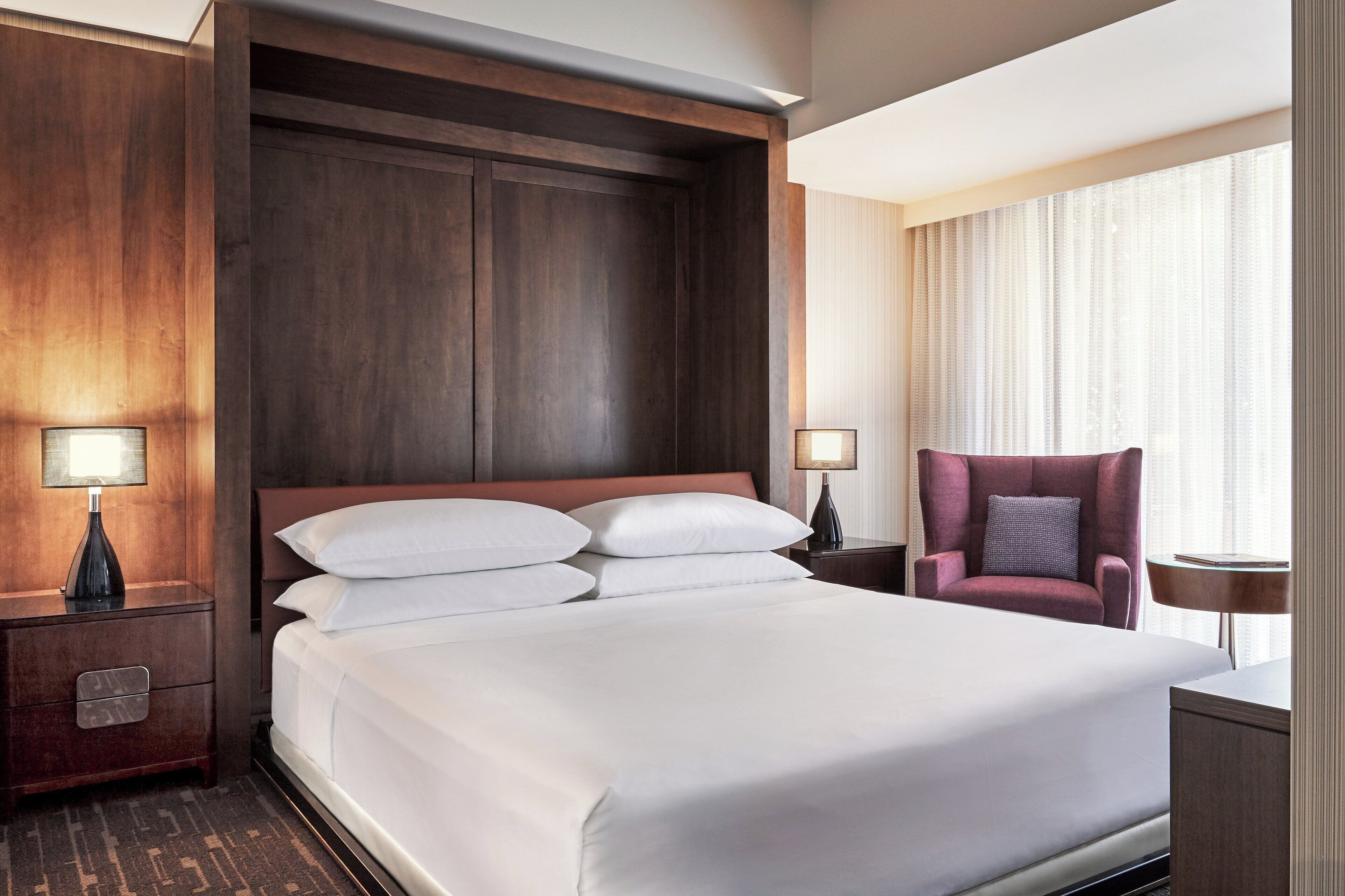 Een hotelkamer met een groot bed, een houten hoofdbord, twee nachtkastjes met lampen, een stoel en een raam met gordijnen.