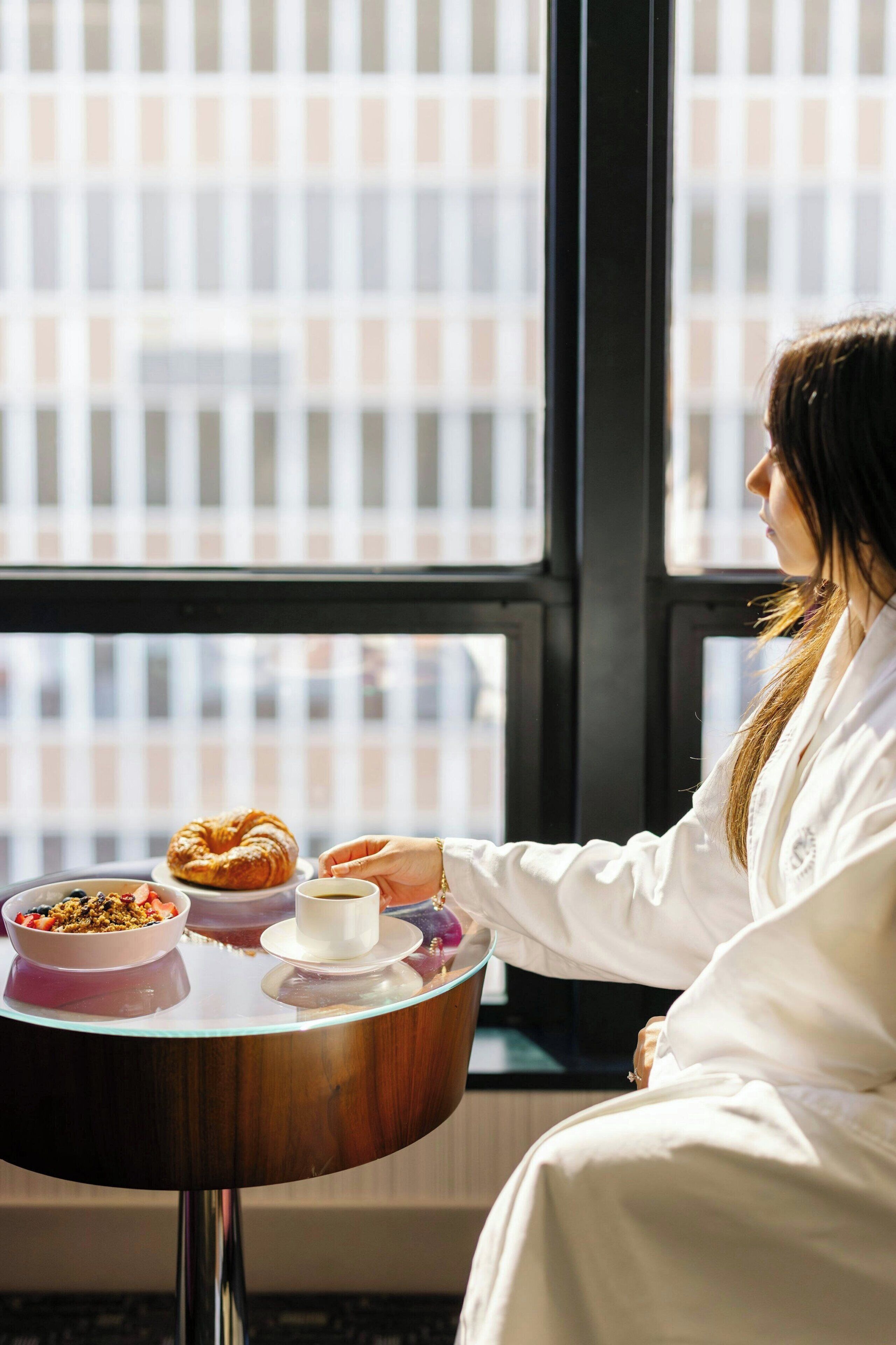 Une personne est assise à une table devant un croissant, un bol de céréales et une tasse de café, et peut admirer la vue sur la ville à travers une fenêtre.