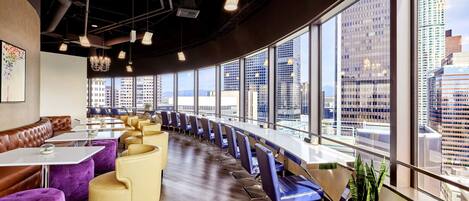 A modern restaurant with a long counter, purple and yellow seating, and large windows offering city views.