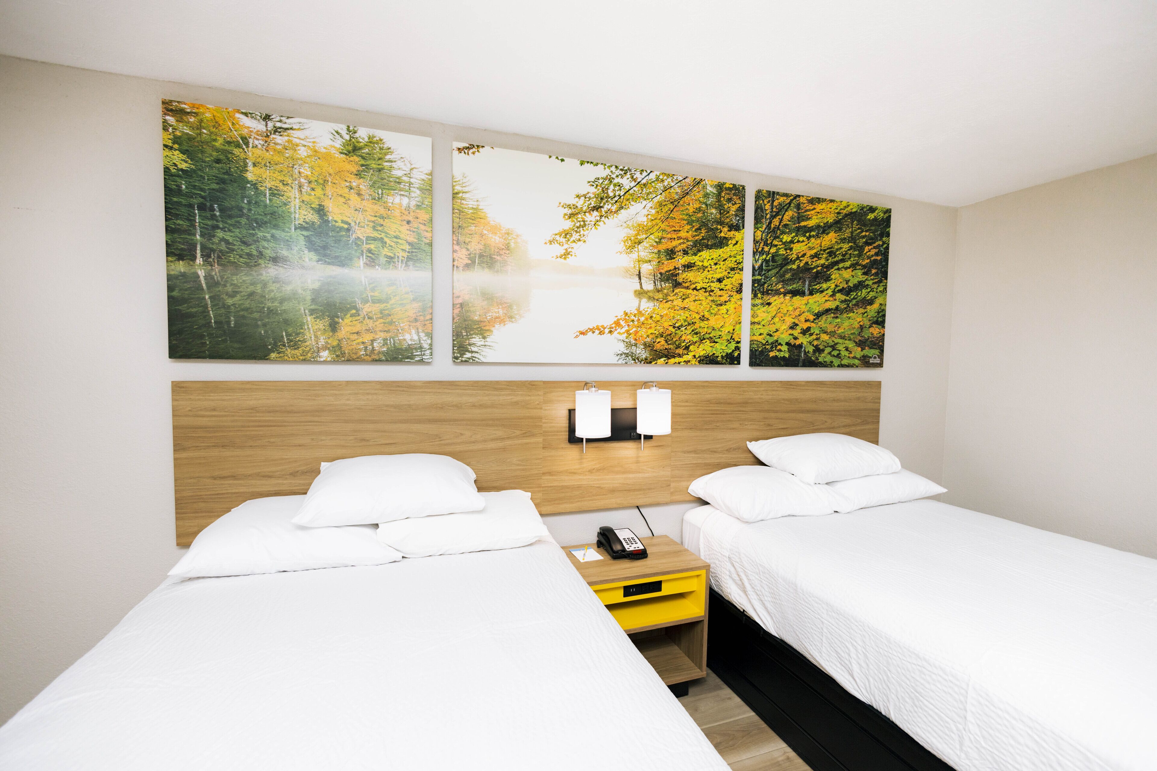 A hotel room with two beds, a wooden headboard, a nightstand with a phone, and a wall-mounted light fixture.