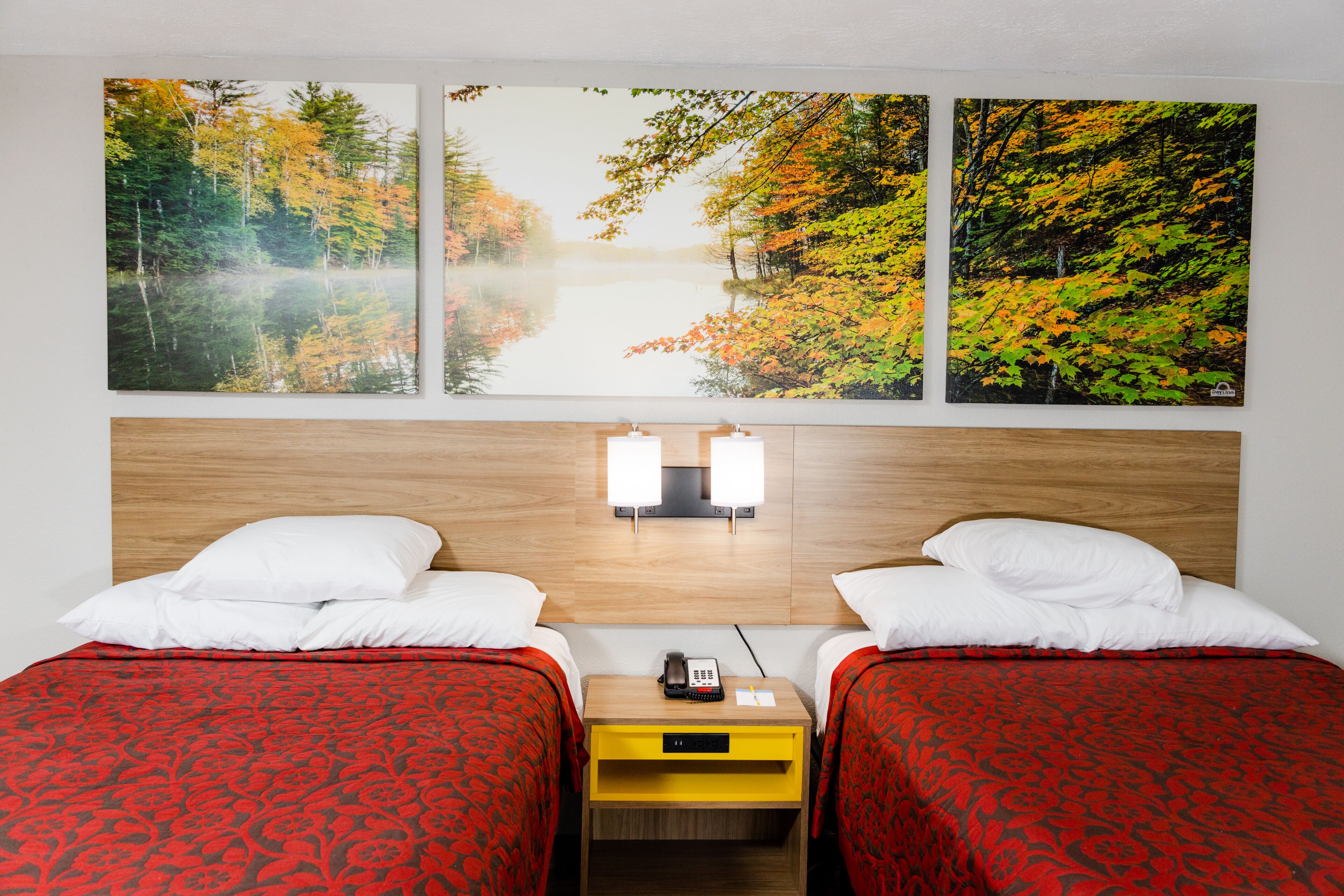 Ein Hotelzimmer mit zwei Betten, einem Holz-Kopfteil, einem Nachttisch mit Telefon und drei großen Herbstwald-Gemälden an der Wand.