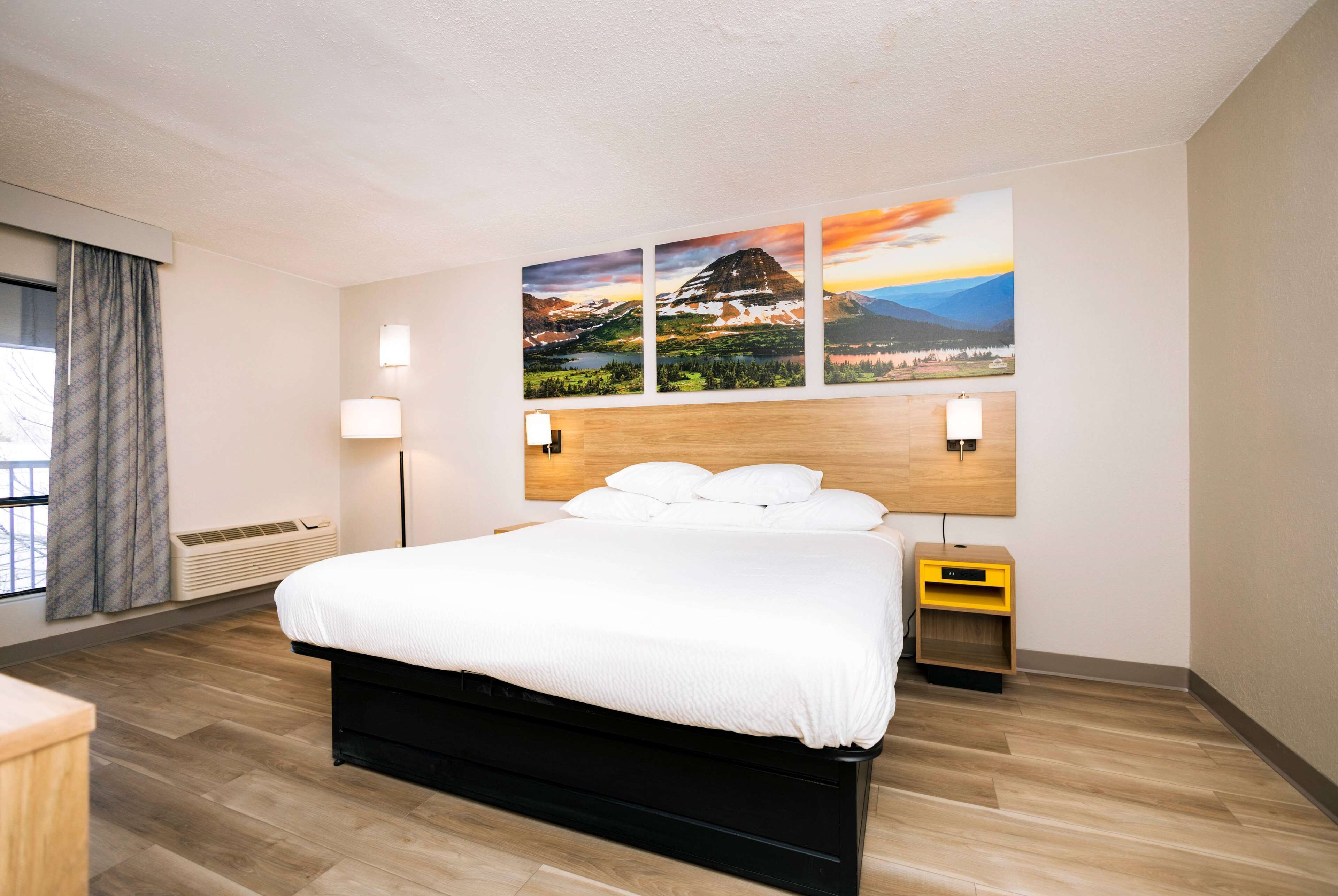 A modern hotel room with a large bed, a wooden headboard, and a wall-mounted painting.