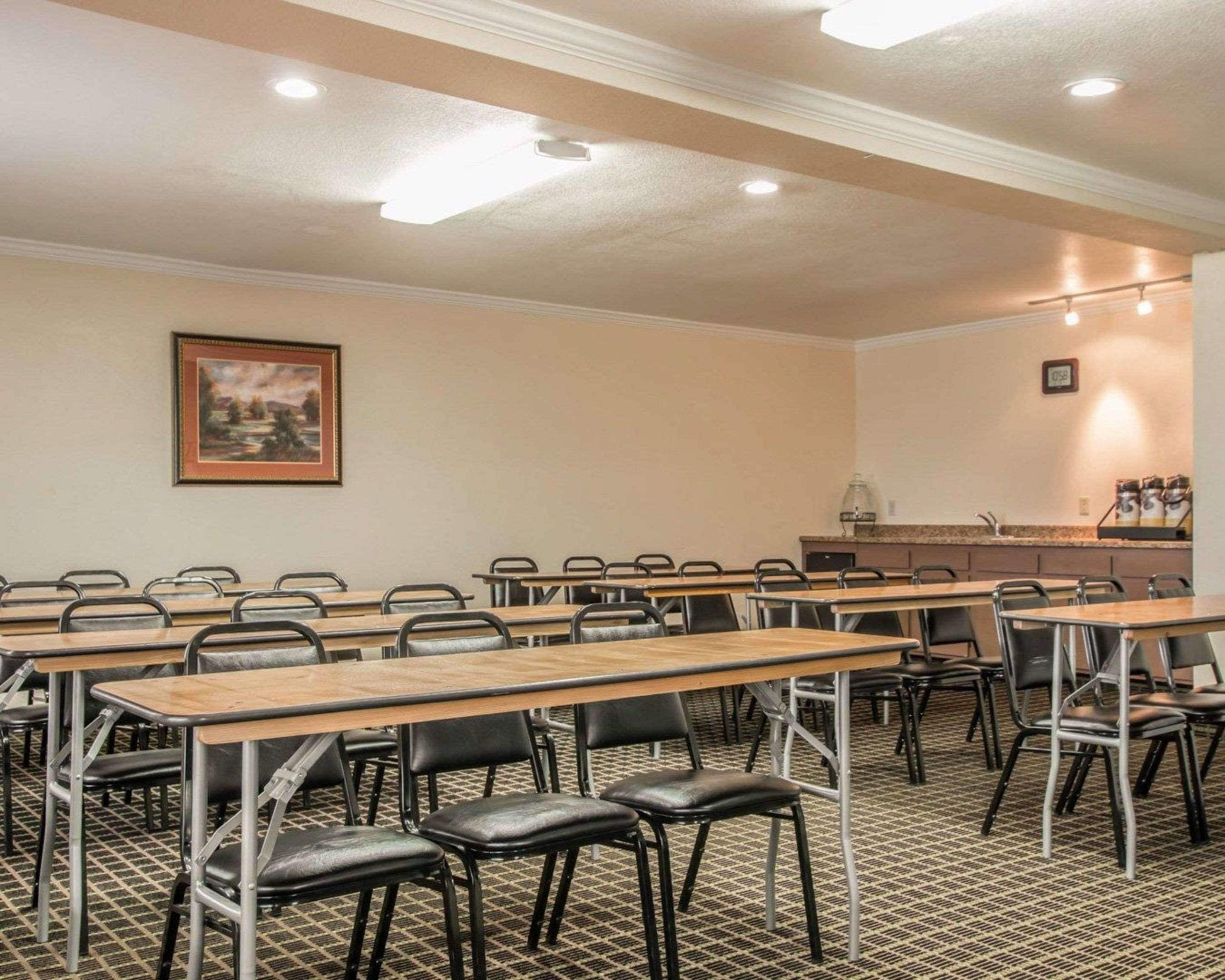 Uma sala de conferências com mesas e cadeiras compridas, um quadro na parede e uma área de cozinha.