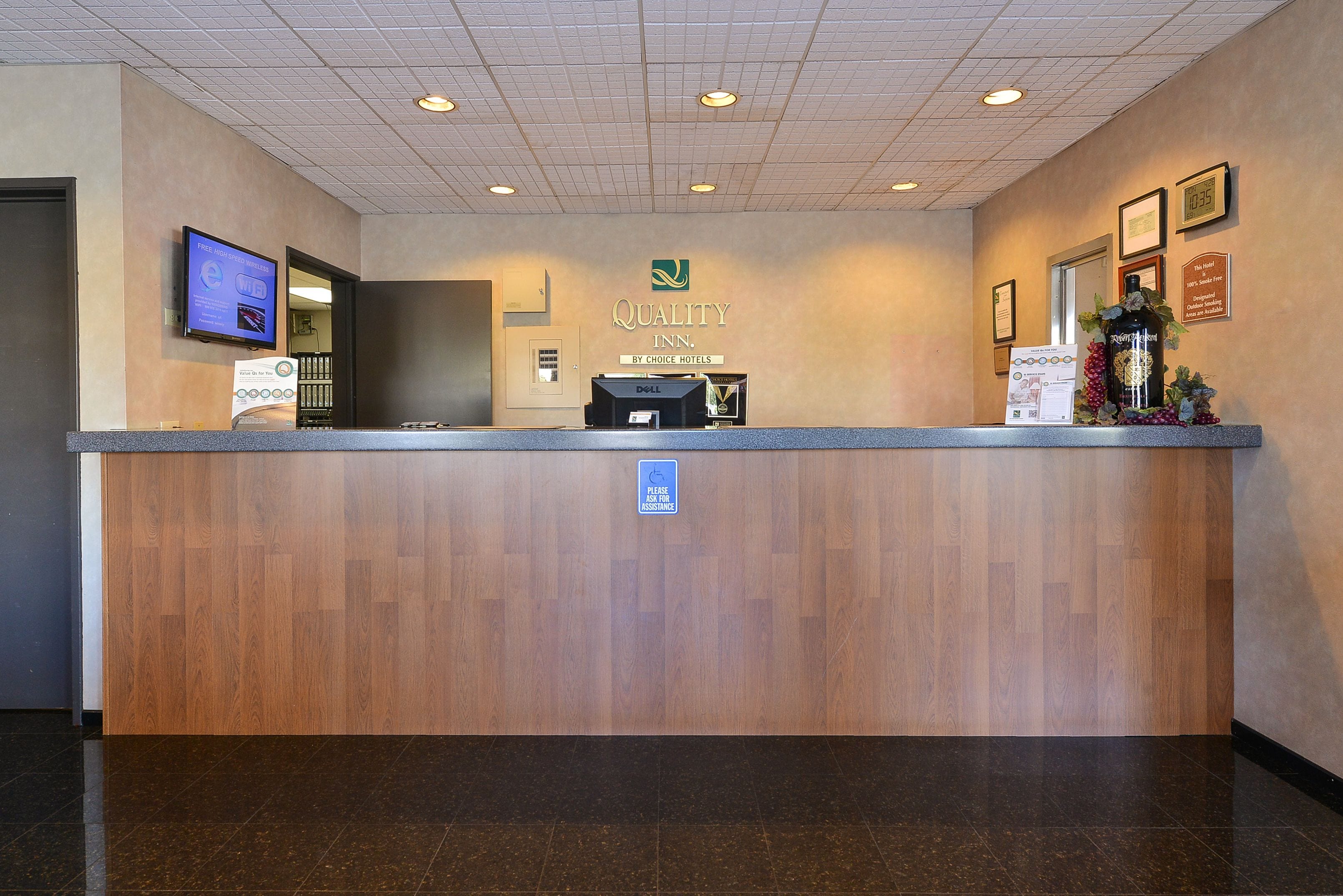 Un mostrador de recepción de hotel con encimera de madera, una pantalla digital y un cartel que indica servicio de atención las 24 horas.