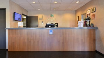 Un mostrador de recepción de hotel con encimera de madera, una pantalla digital y un cartel que indica servicio de atención las 24 horas.