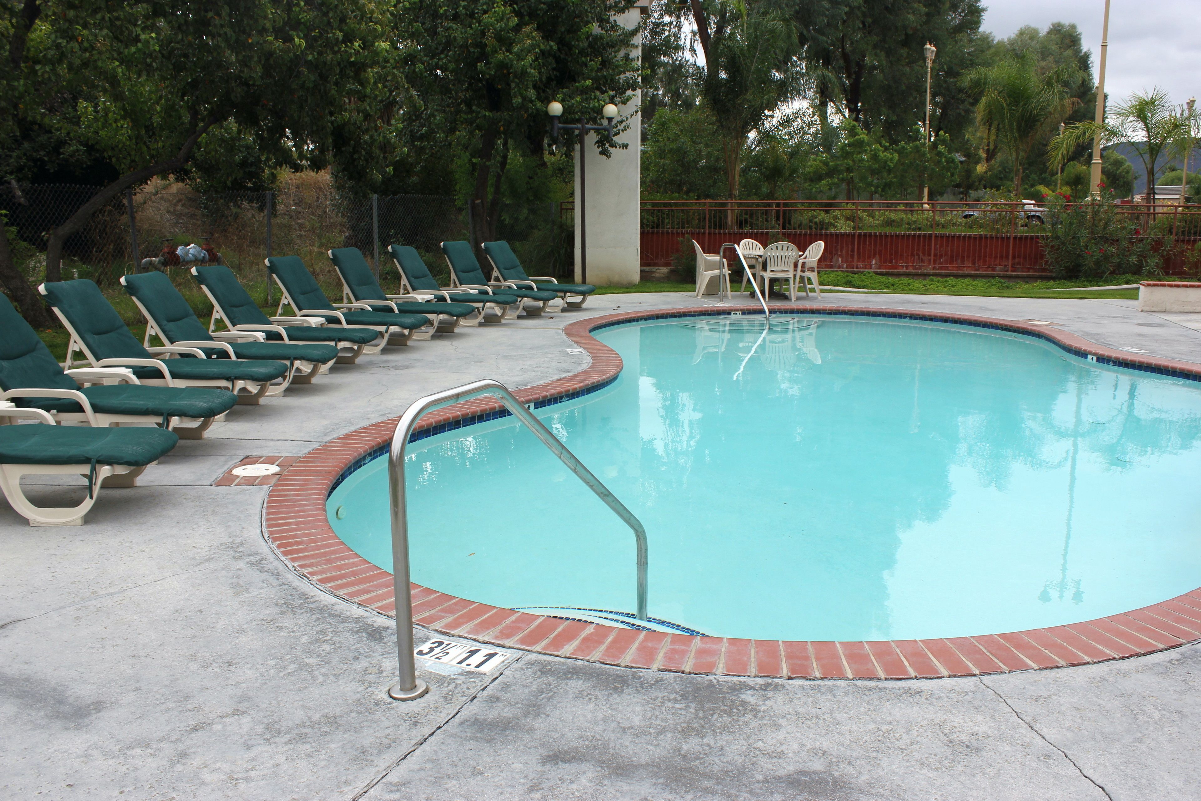 Piscina con borde de ladrillo, rodeada de sillas reclinables verdes y una pequeña mesa con sillas.
