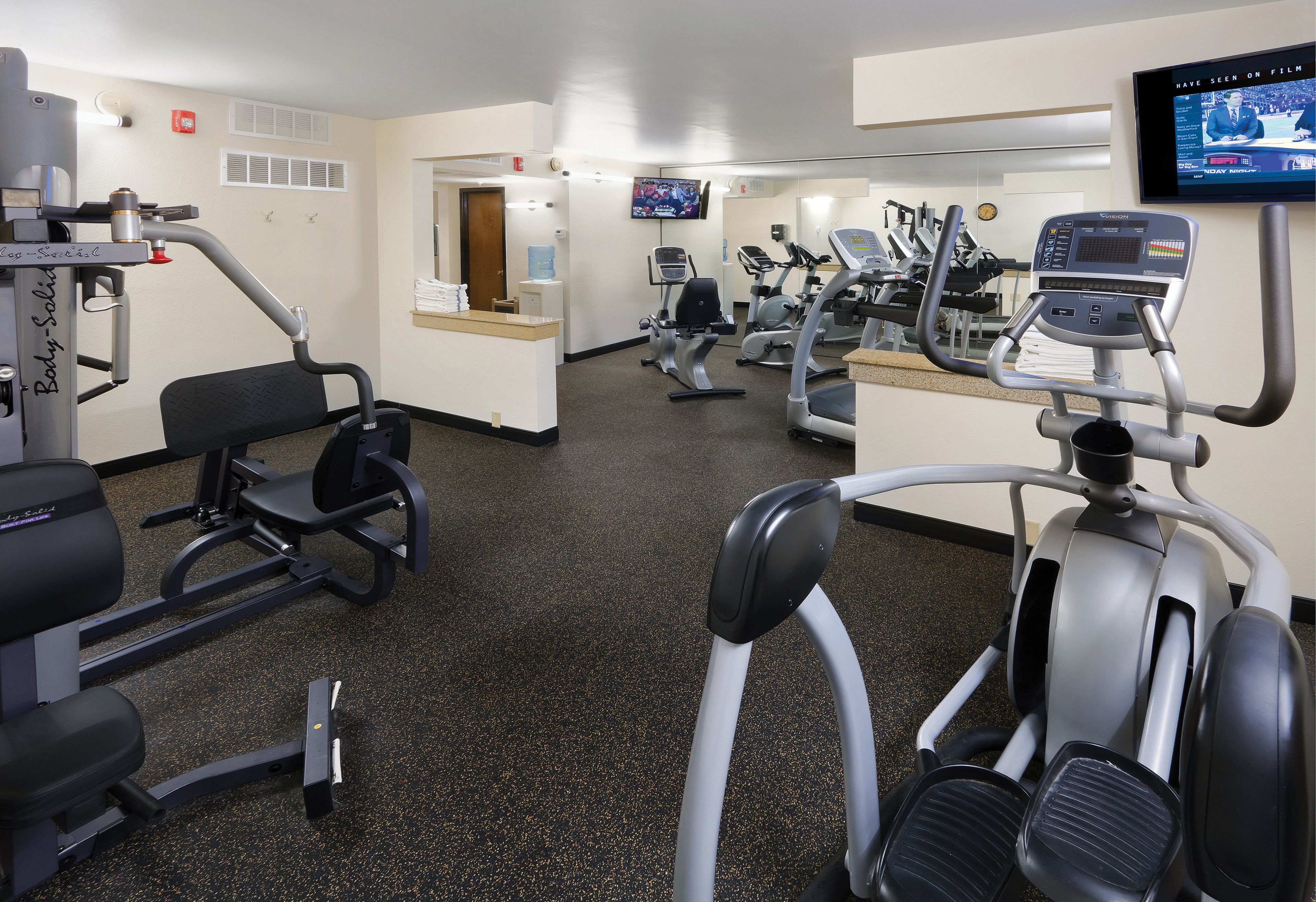 Un gimnasio con máquinas de cardio, máquinas de pesas y una pantalla de televisión mostrando un noticiero.