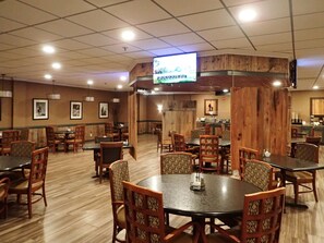 A restaurant with multiple dining tables and chairs, a television mounted on the ceiling, and framed pictures on the walls. - Western Heritage Hotel and Conference Center, an Ascend Collection Hotel (Casper)