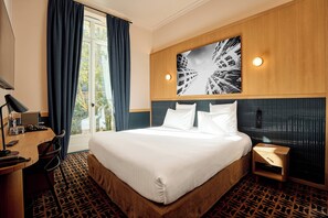 Habitación de hotel con una cama grande, un escritorio con lámpara, una silla, una mesita y un ventanal con cortinas.