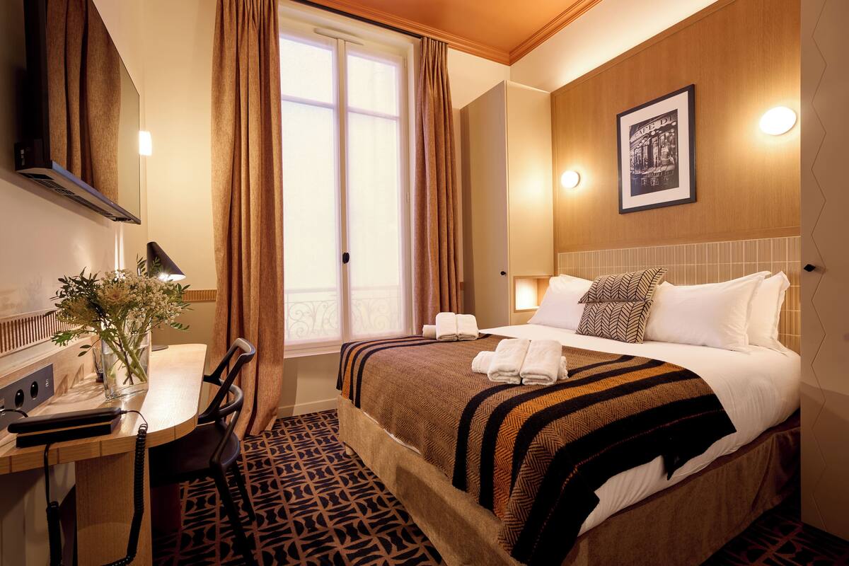A hotel room with a bed, a desk with a lamp, a chair, a window with curtains, and a framed picture on the wall.