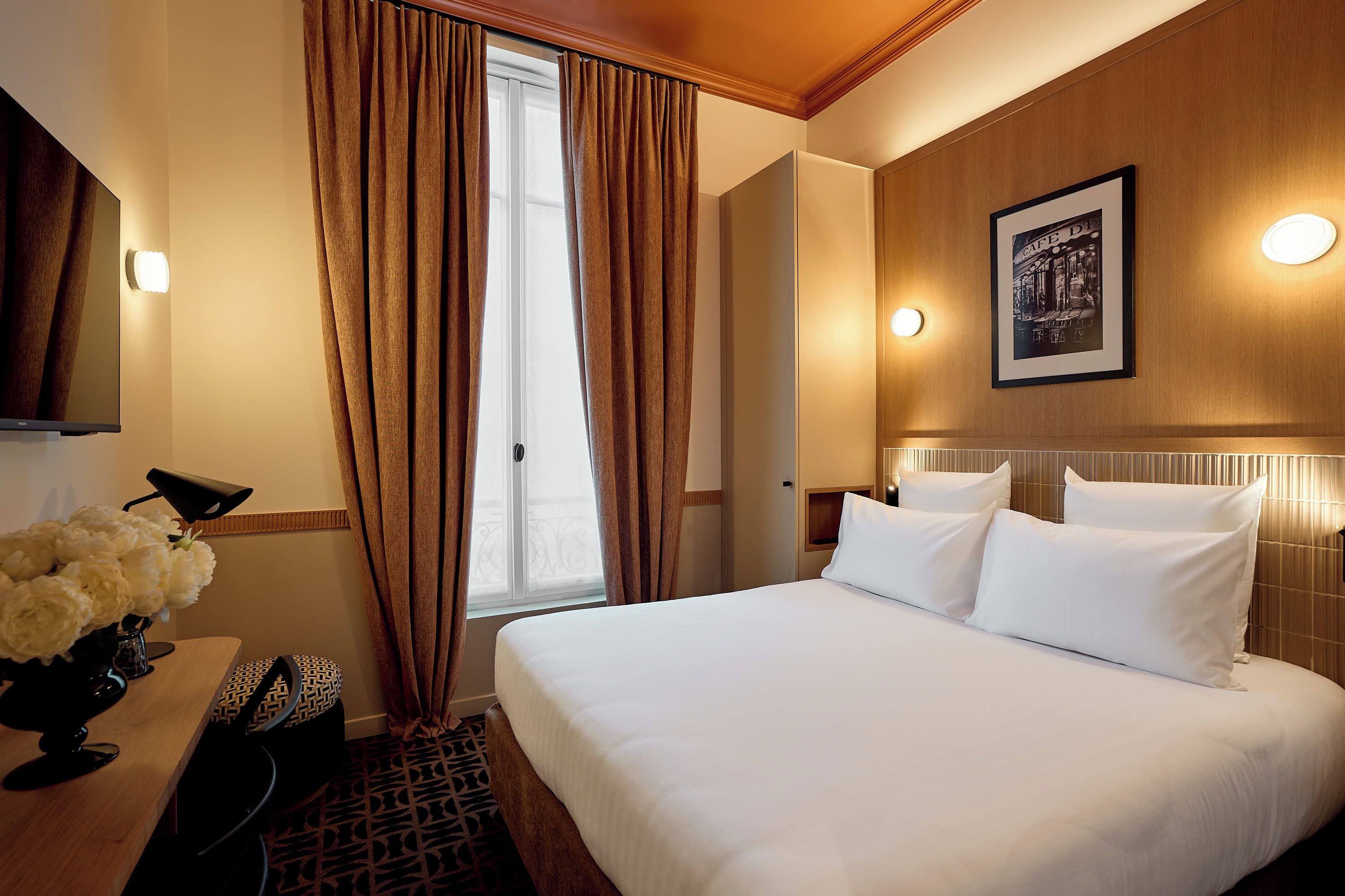 A hotel room with a bed, a desk with a lamp, a vase of flowers, a framed picture on the wall, and a window with curtains.