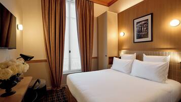 A hotel room with a bed, a desk with a lamp, a vase of flowers, a framed picture on the wall, and a window with curtains.