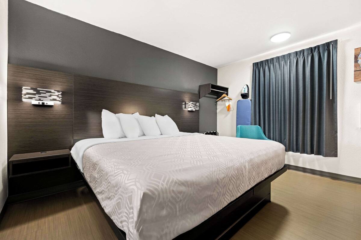A modern hotel room with a large bed, dark wood paneling, a built-in shelf, and a window with curtains.