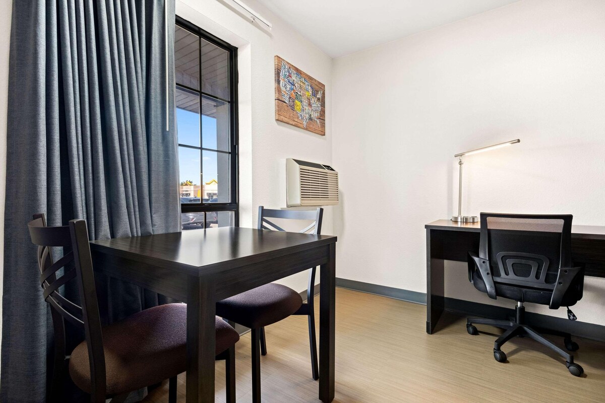 Una habitación con una mesa de madera, dos sillas, un escritorio con silla, una ventana con cortinas y un aire acondicionado instalado en la pared.