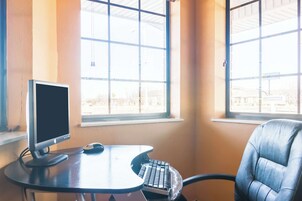 A computer desk with a monitor, keyboard, and mouse in a room with large windows and a chair.