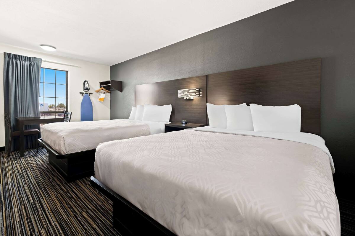 A hotel room with two beds, a desk, a chair, a large window with curtains, and a modern wall design.