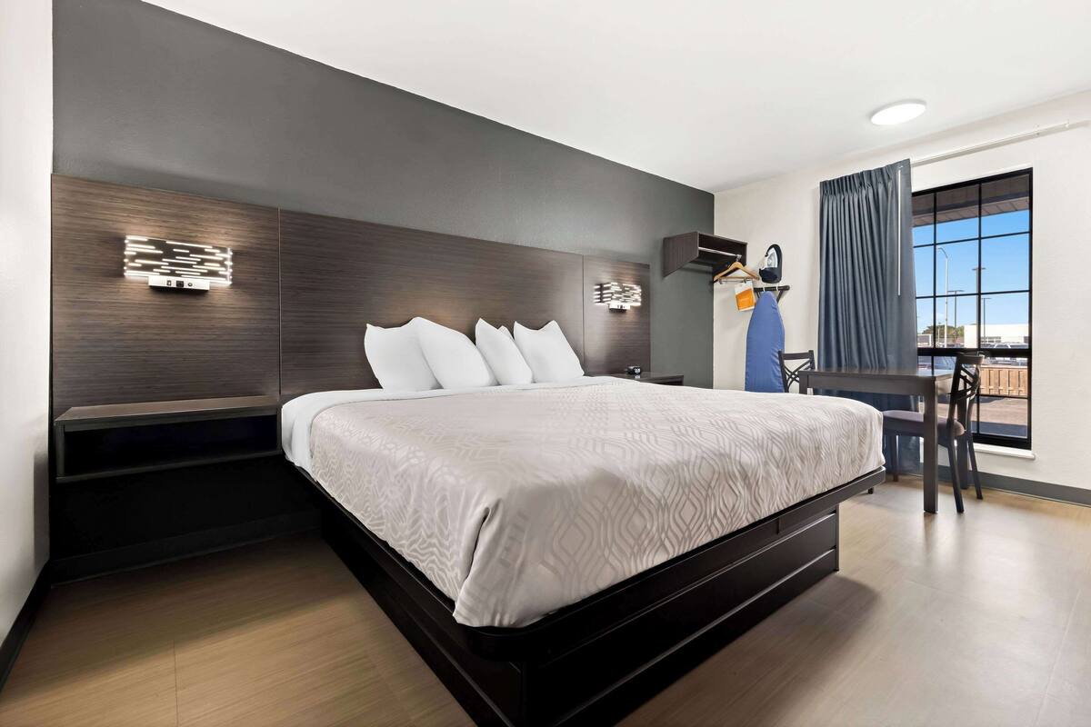 A modern hotel room with a large bed, built-in bedside tables, a desk with a chair, and a large window offering a view.