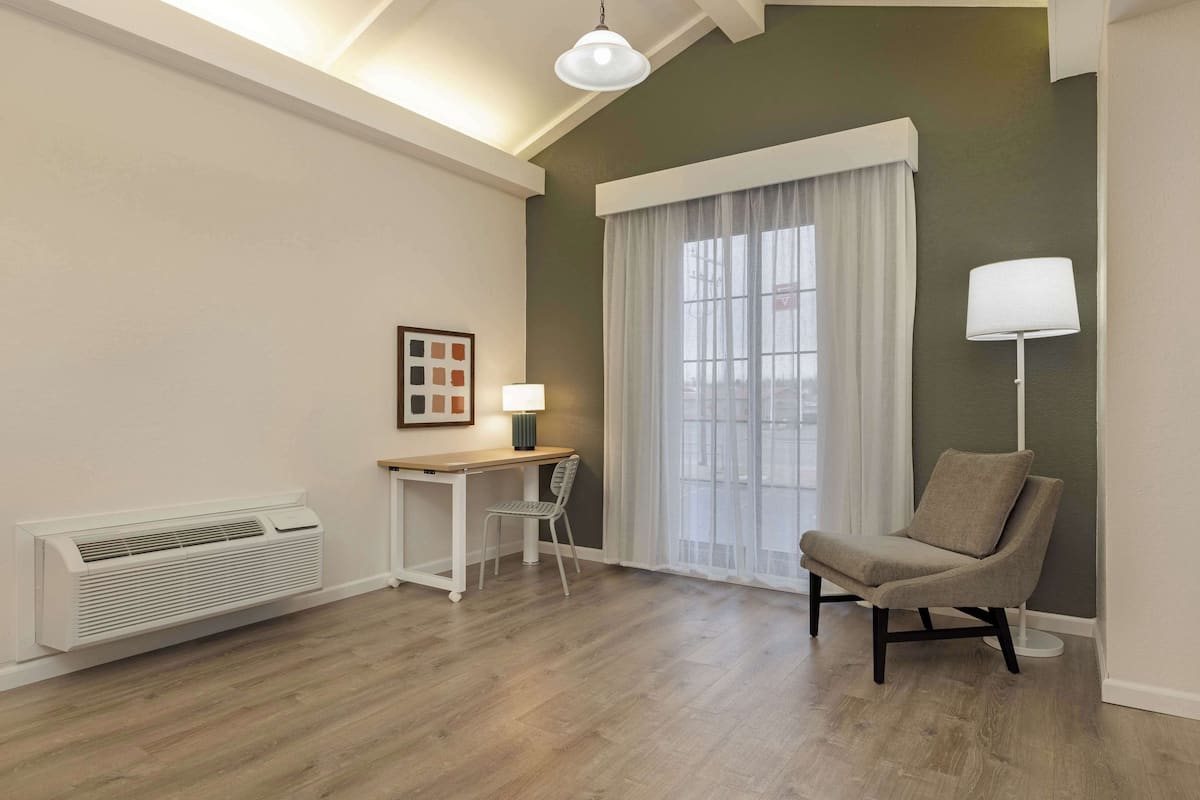 A modern room with a wooden floor, a desk with a chair, a lamp, a radiator, and a large window with curtains.