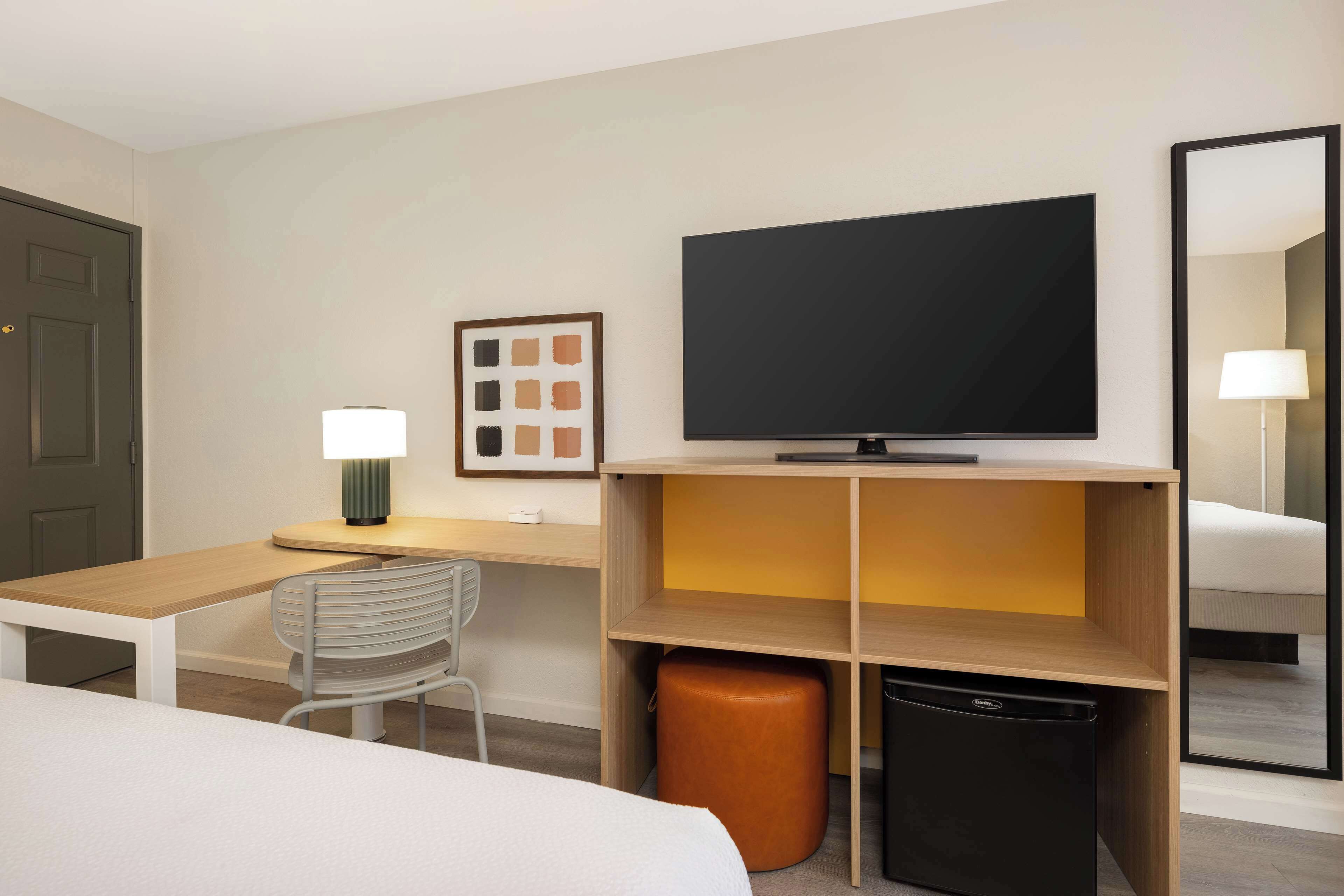 Une chambre d’hôtel comprenant un lit, un bureau avec une lampe, une télévision, une chaise, un petit tabouret et une poubelle.
