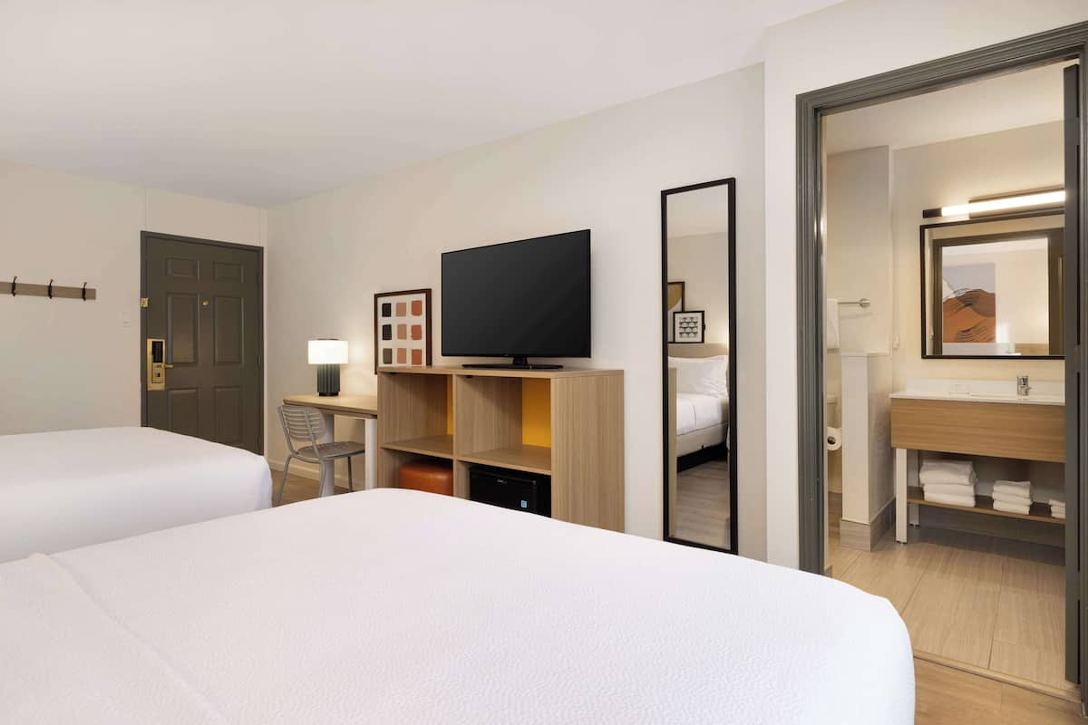 A hotel room with two beds, a desk with a flat-screen TV, a mirror, and a bathroom with a sink and towel rack.