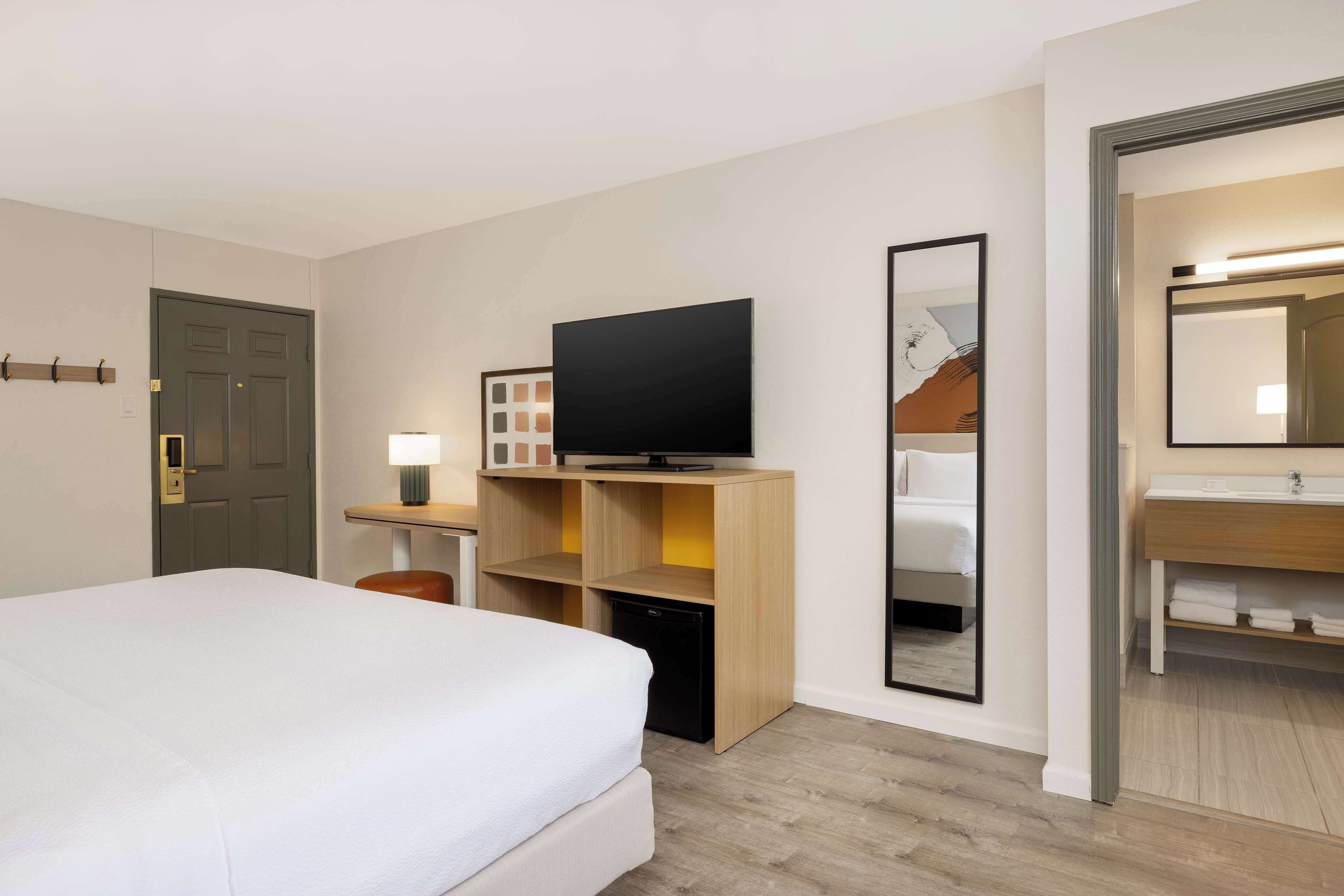 A hotel room with a bed, a TV on a wooden shelf, a desk with a lamp, a mirror, and a bathroom with a sink and towel rack.