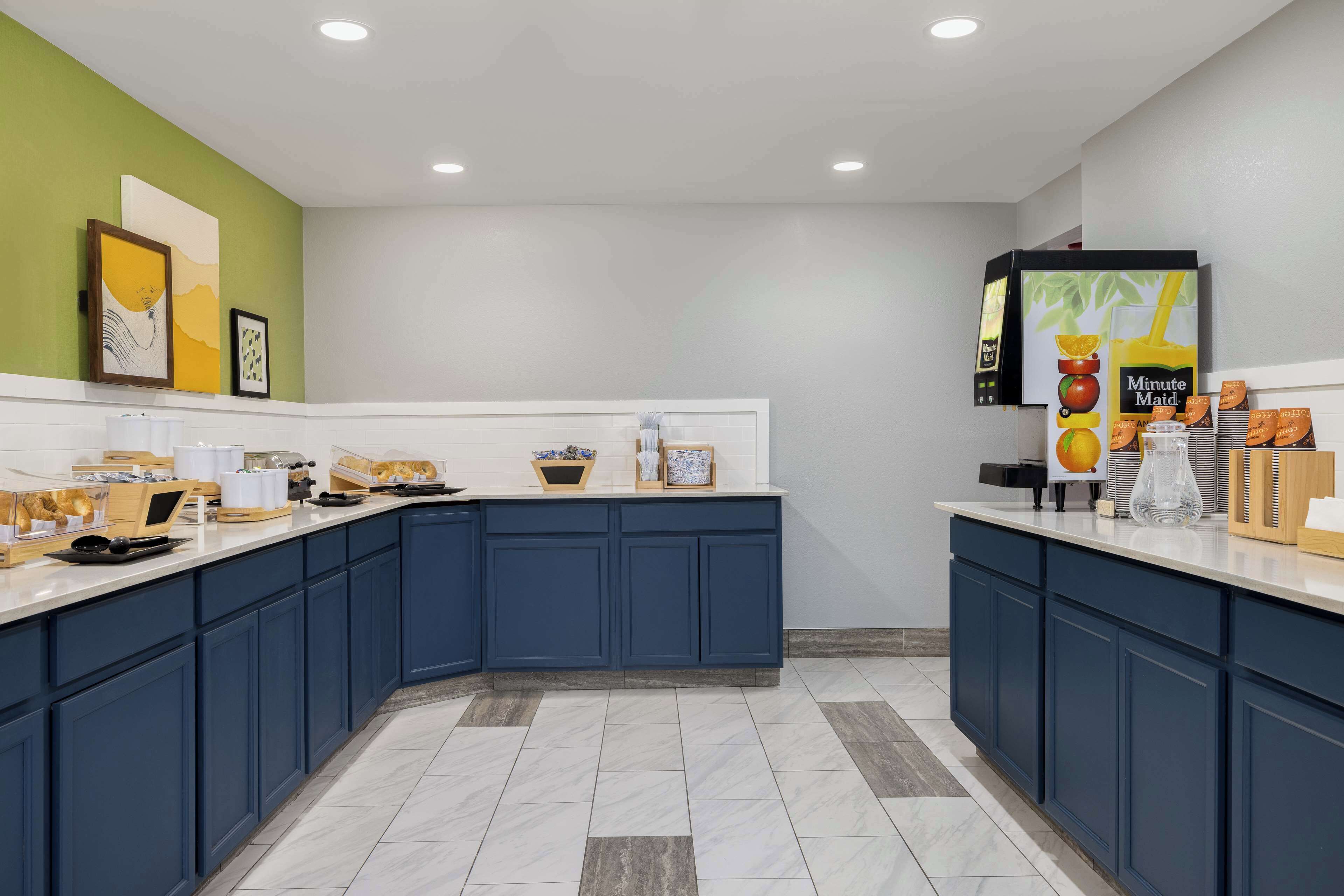 Une cuisine moderne avec des meubles de couleur bleu foncé, un plan de travail blanc et un mur d’accent vert. On y trouve des tableaux encadrés, une machine à café et un distributeur de jus.