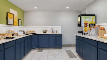 Una cocina moderna con armarios azul oscuro, encimera blanca y una pared de acento verde. Hay cuadros enmarcados, una máquina de café y un dispensador de zumo.
