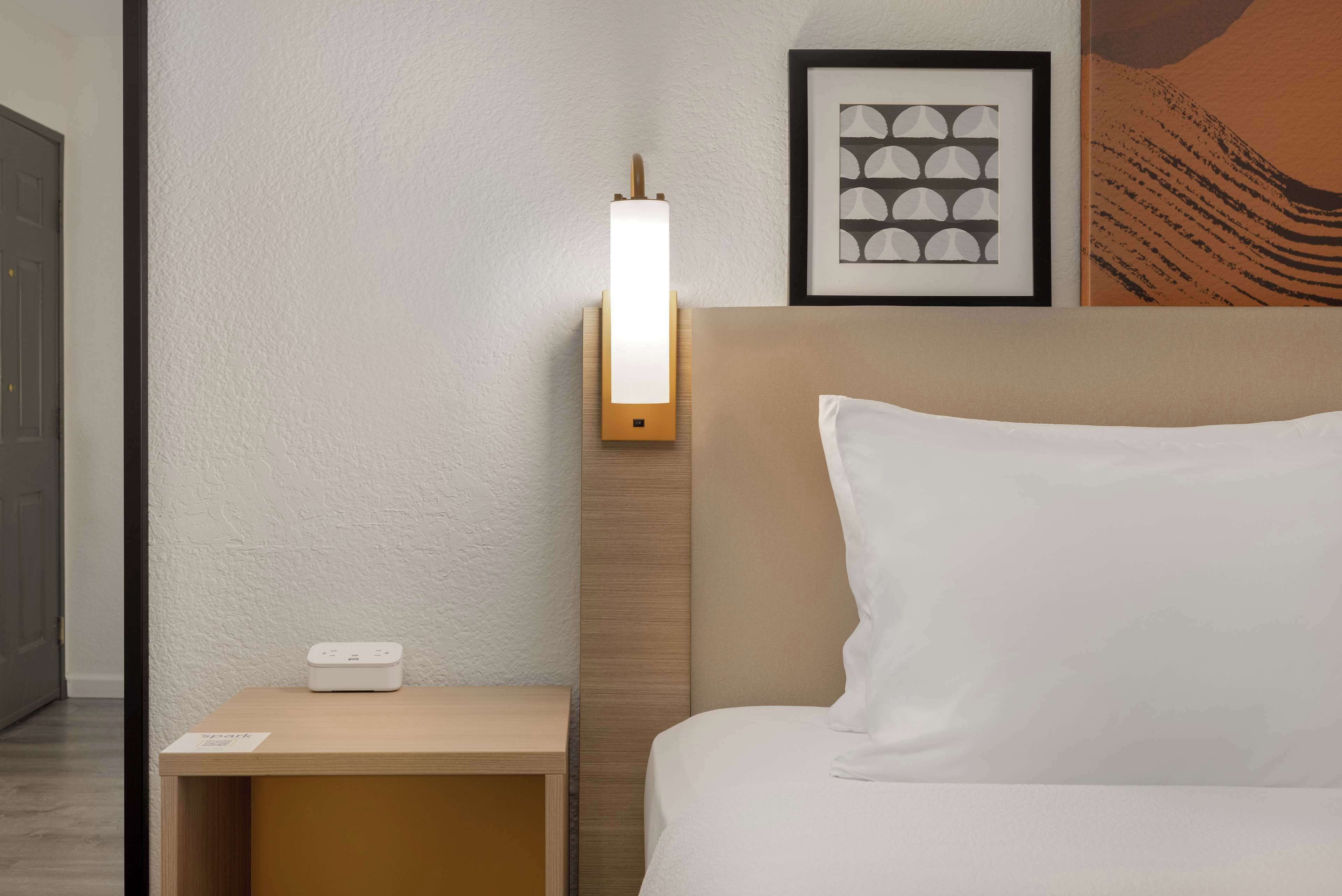 A hotel room with a bedside table, a wall-mounted light, a framed artwork, and a bed with white bedding.