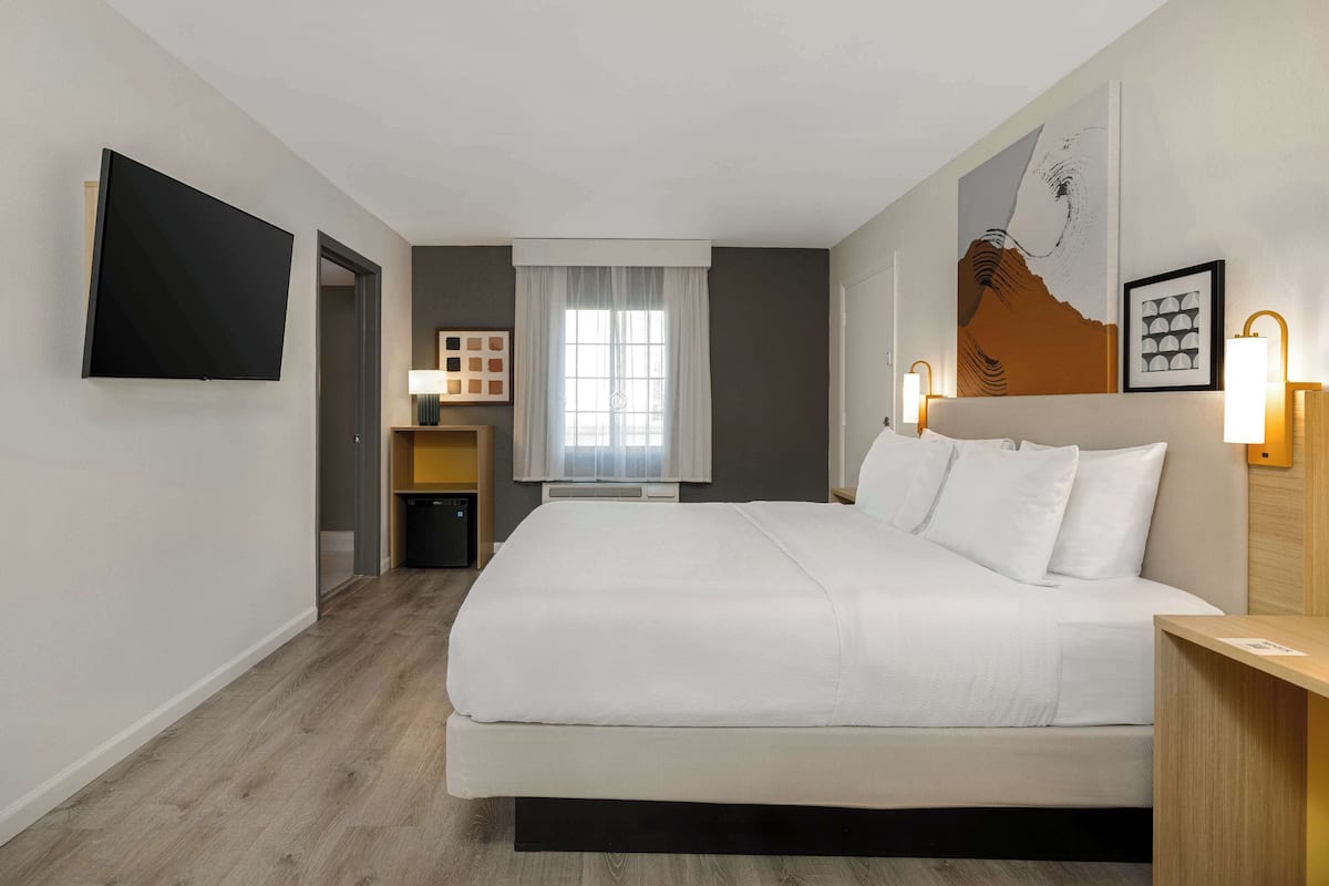 A hotel room with a large bed, a flat-screen TV mounted on the wall, a window with curtains, and a wall-mounted lamp.