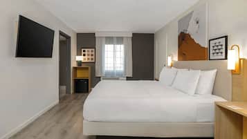 A hotel room with a large bed, a flat-screen TV mounted on the wall, a window with curtains, and a wall-mounted lamp.
