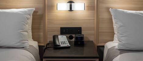 A hotel room with two beds, a bedside table with a phone and a digital clock, and a wall-mounted light fixture.