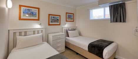 Une chambre avec deux lits simples, une table de chevet avec une lampe, une commode et trois tableaux encadrés accrochés au mur.