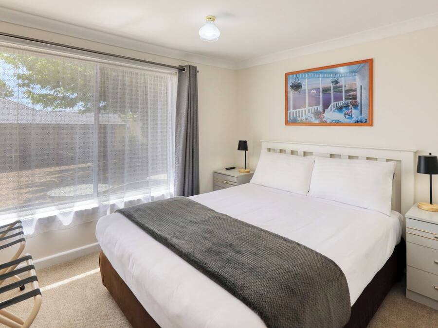 A hotel room with a large bed, a bedside table with a lamp, a chair, a window with curtains, and a painting on the wall.