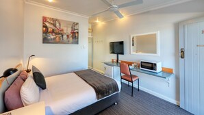 Una habitación de hotel con una cama, un escritorio con computadora, un televisor, un microondas y un cuadro de un paisaje urbano.