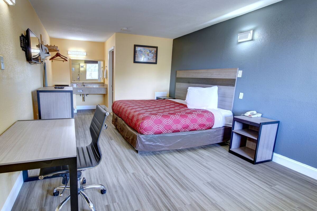 A hotel room with a bed, a desk, a chair, a small table, a mirror, and a nightstand.
