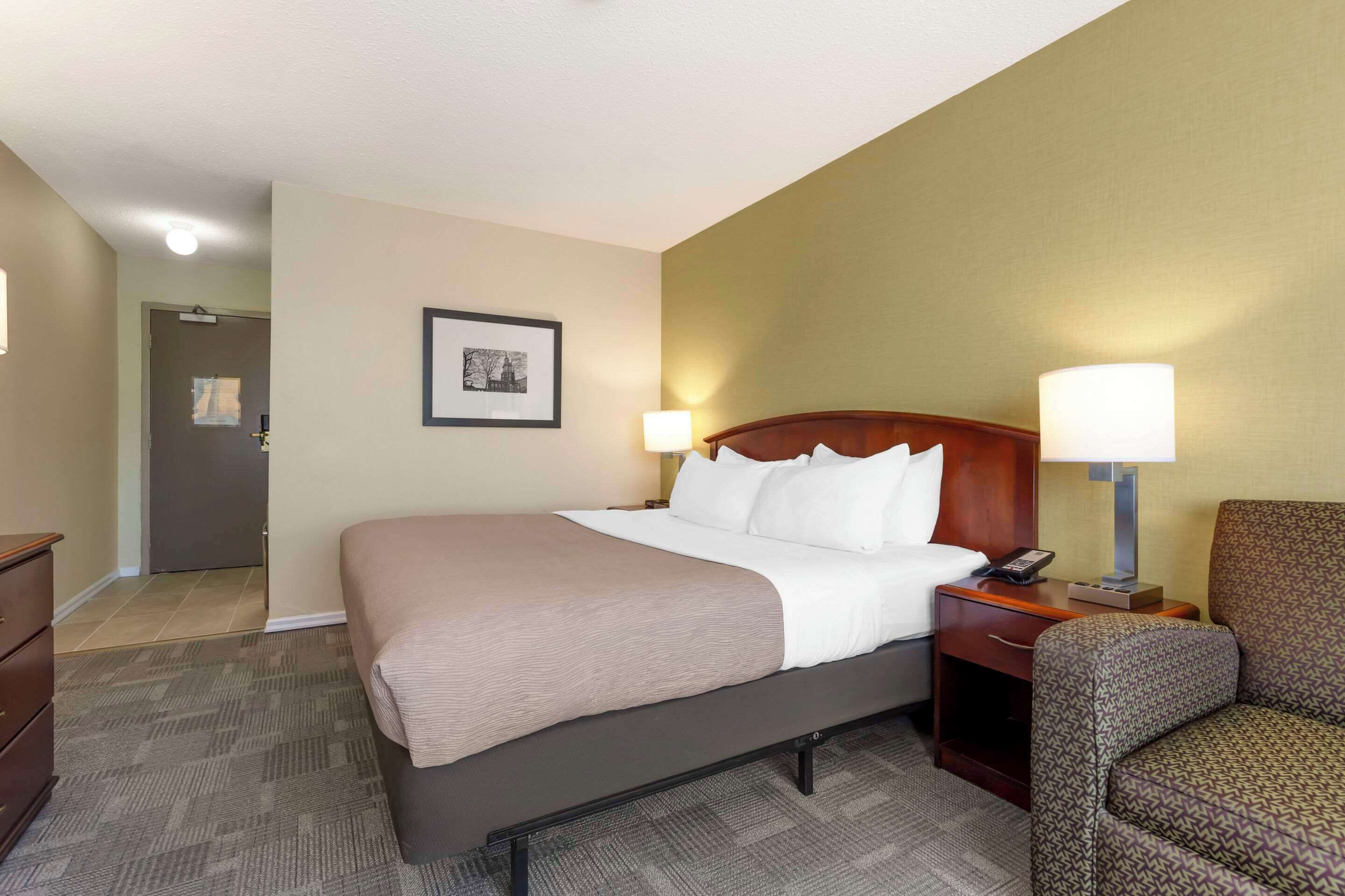 A hotel room with a large bed, two bedside lamps, a wooden headboard, a framed picture on the wall, and a chair.