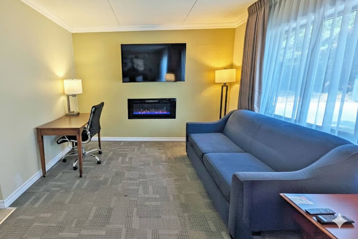 A hotel room with a sofa, a desk with a chair, a flat-screen TV mounted on the wall, and a fireplace.
