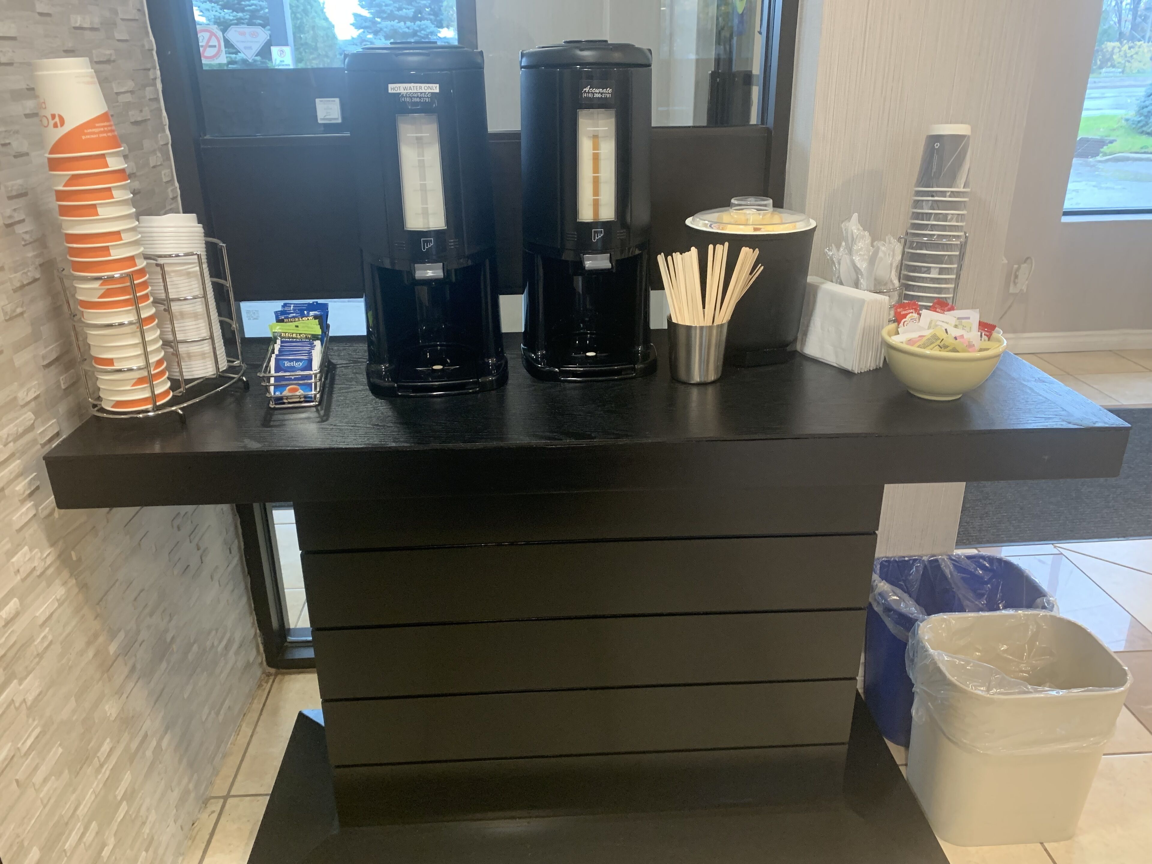 A coffee station with two coffee makers, a stack of disposable cups, a bowl of straws, and a trash bin.