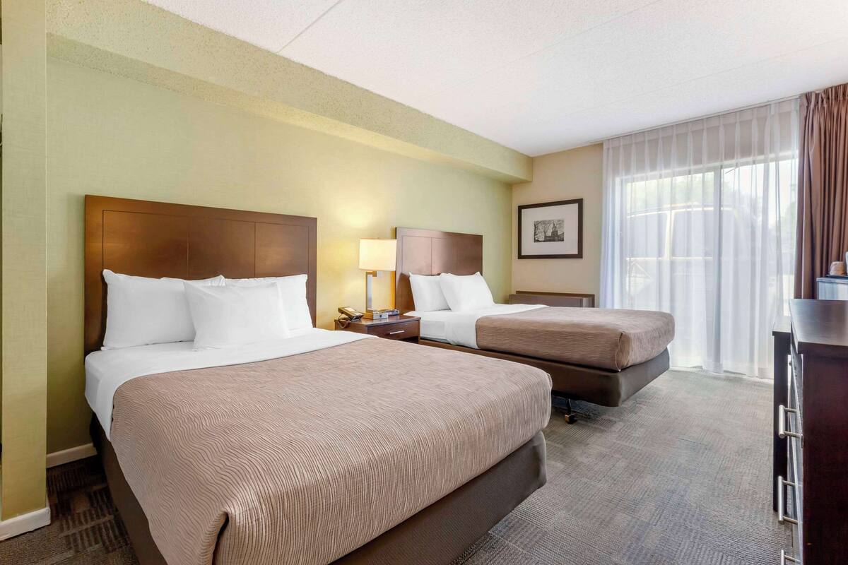 A hotel room with two beds, a desk, a chair, and a large window with curtains.