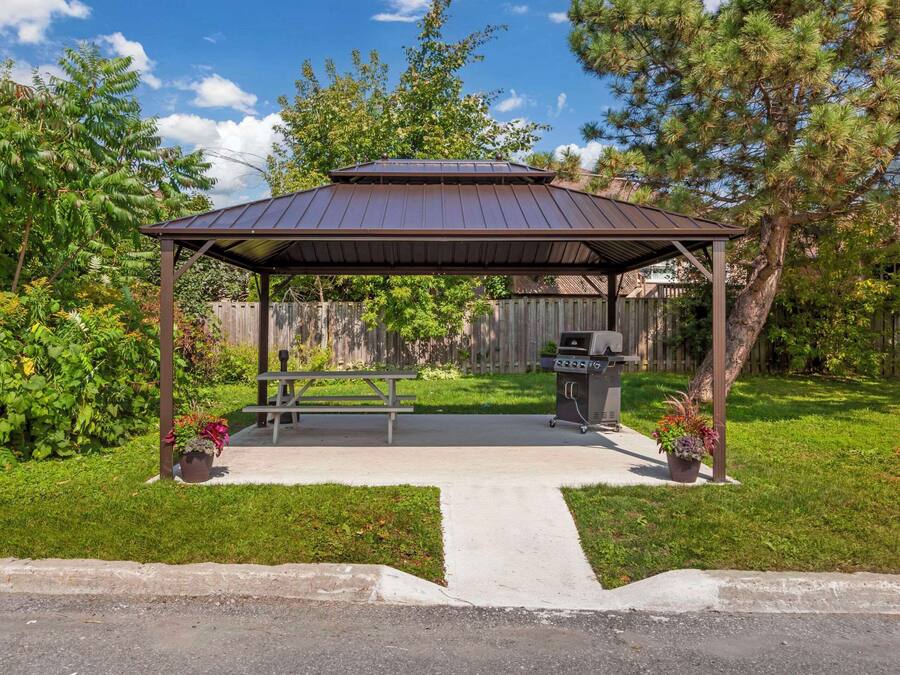A covered outdoor area with a picnic table and grill, surrounded by greenery and a wooden fence.