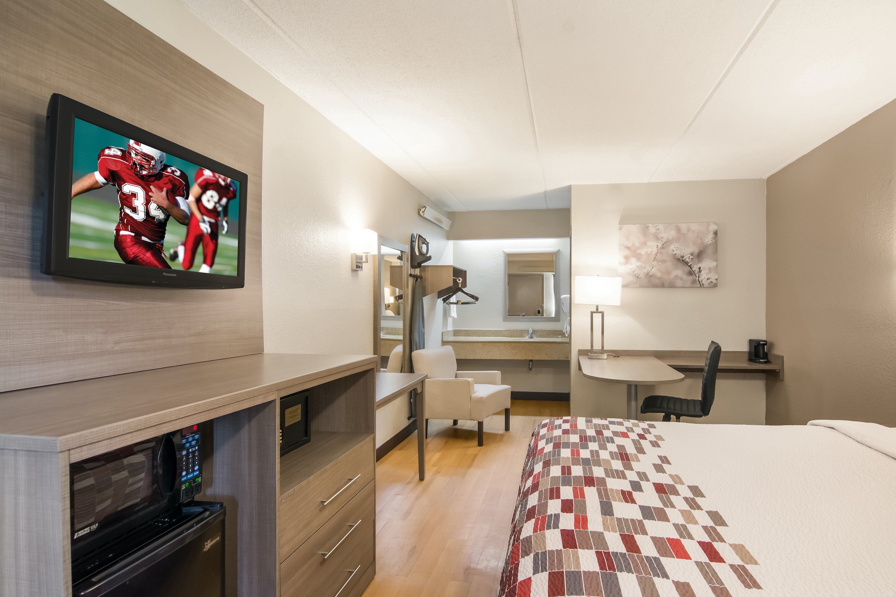 A hotel room with a flat-screen TV, a bed, a desk, a chair, a small table, a mirror, and a coffee machine.