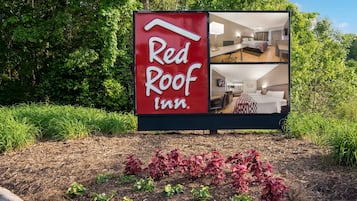 Señal de Red Roof Inn con vistas a una habitación que incluye una cama, un escritorio y una silla.