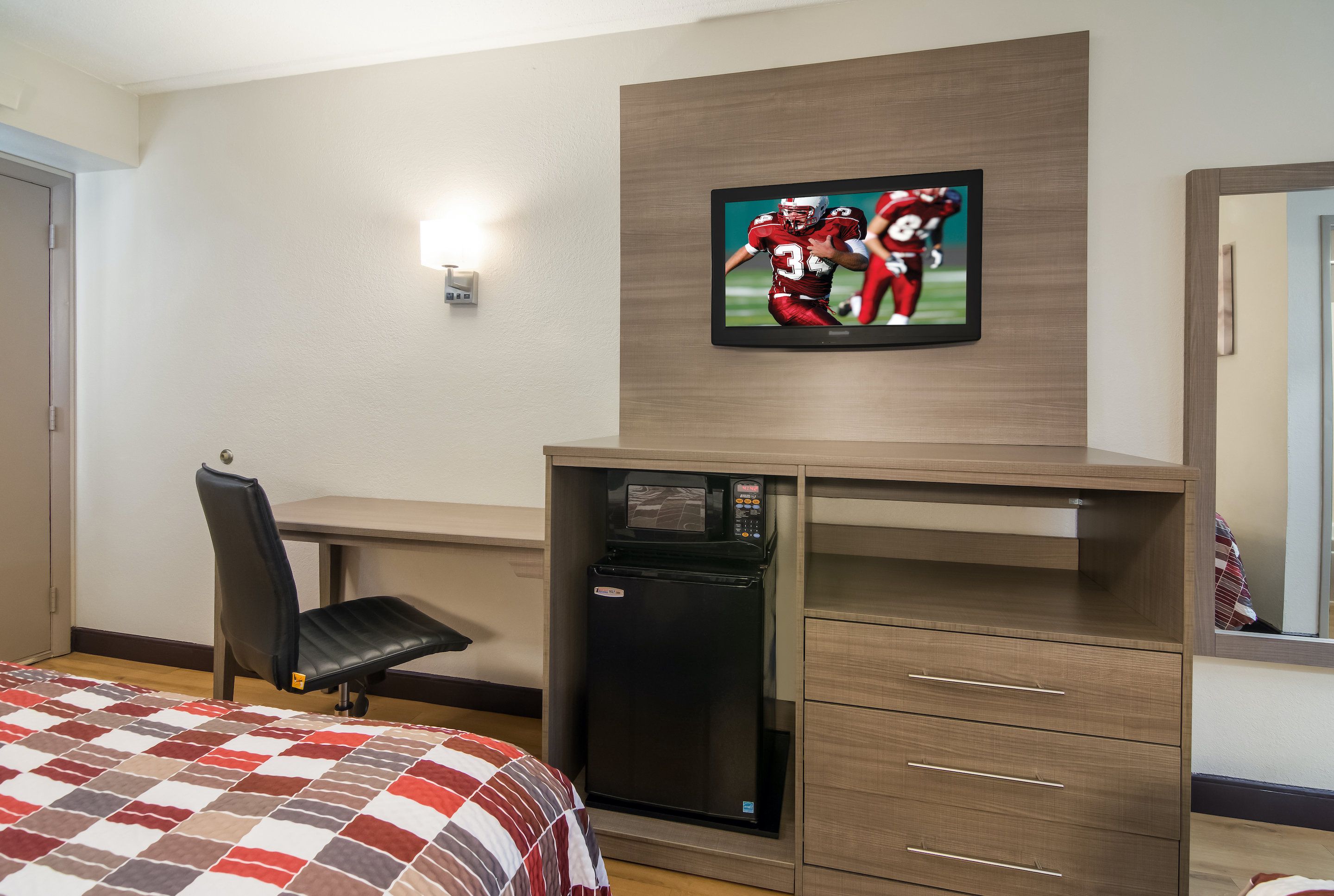 A hotel room with a bed, a desk, a refrigerator, a television mounted on the wall, and a mirror.