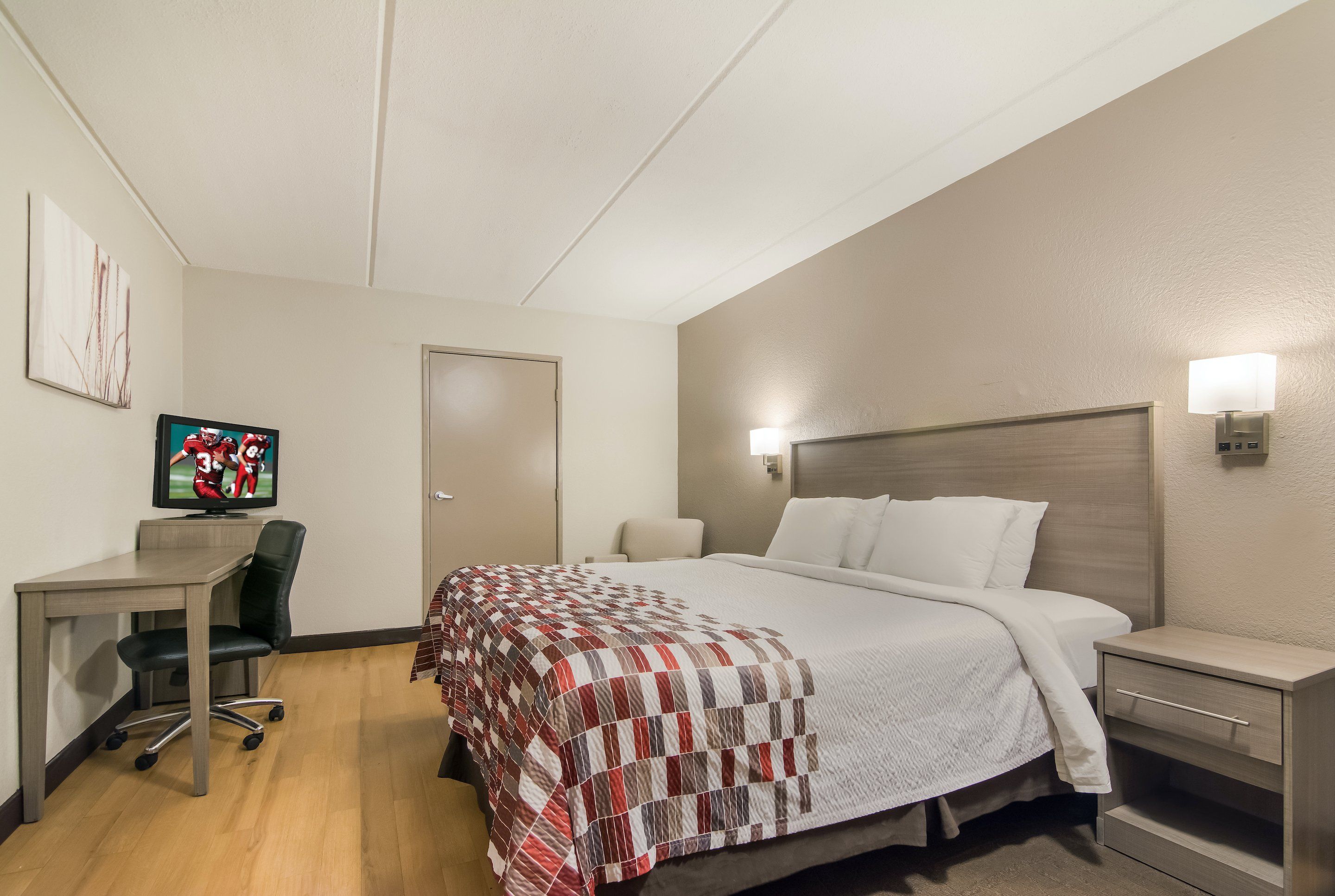 A hotel room with a bed, bedside table, lamp, desk with chair, television, and artwork on the wall.