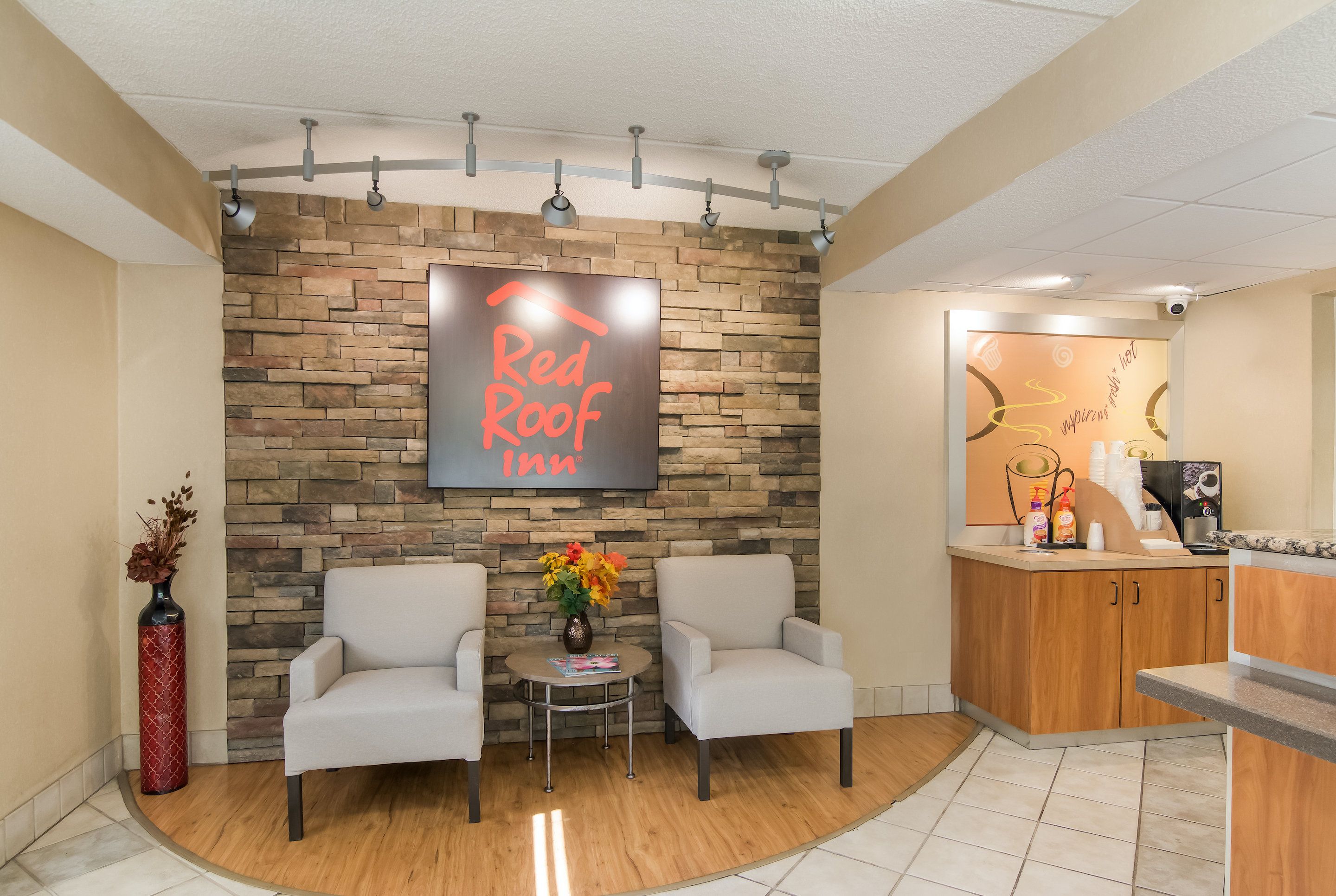 A Red Roof Inn reception area with a stone wall, two armchairs, a small table with a vase, and a wooden counter with amenities.