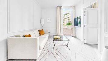 Een moderne hotelkamer met een witte bank, een glazen salontafel en uitzicht op een gebouw door een glazen deur.