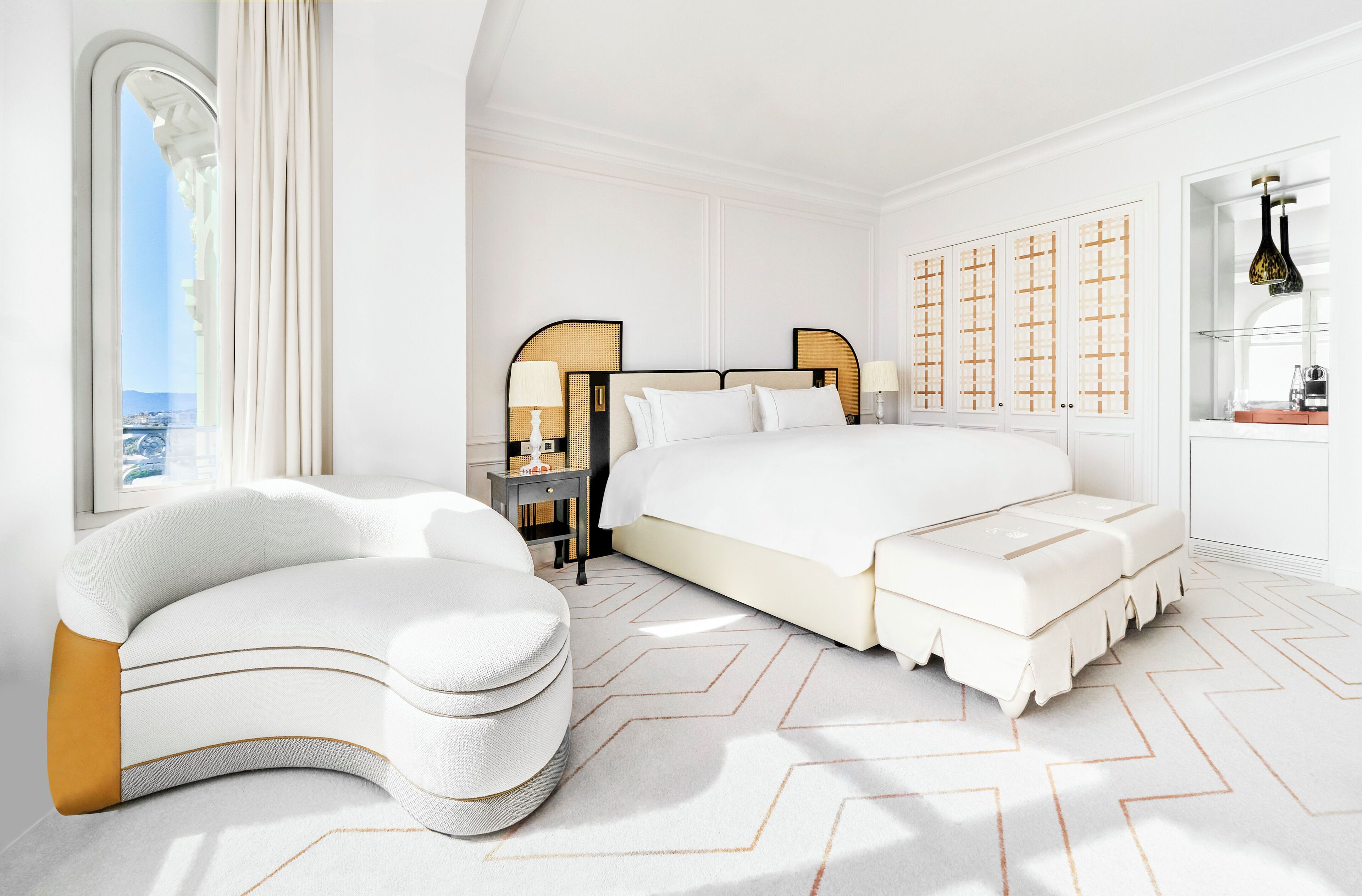 A modern bedroom with a large bed, a curved white sofa, a nightstand, and a wardrobe with a geometric pattern.