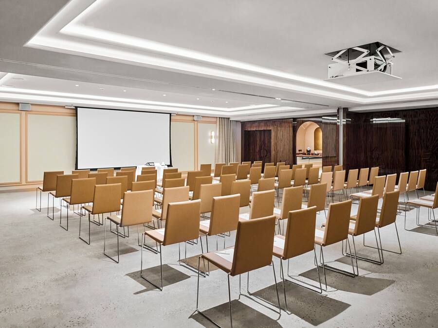 A conference room with rows of chairs, a projection screen, and a wooden panel wall.
