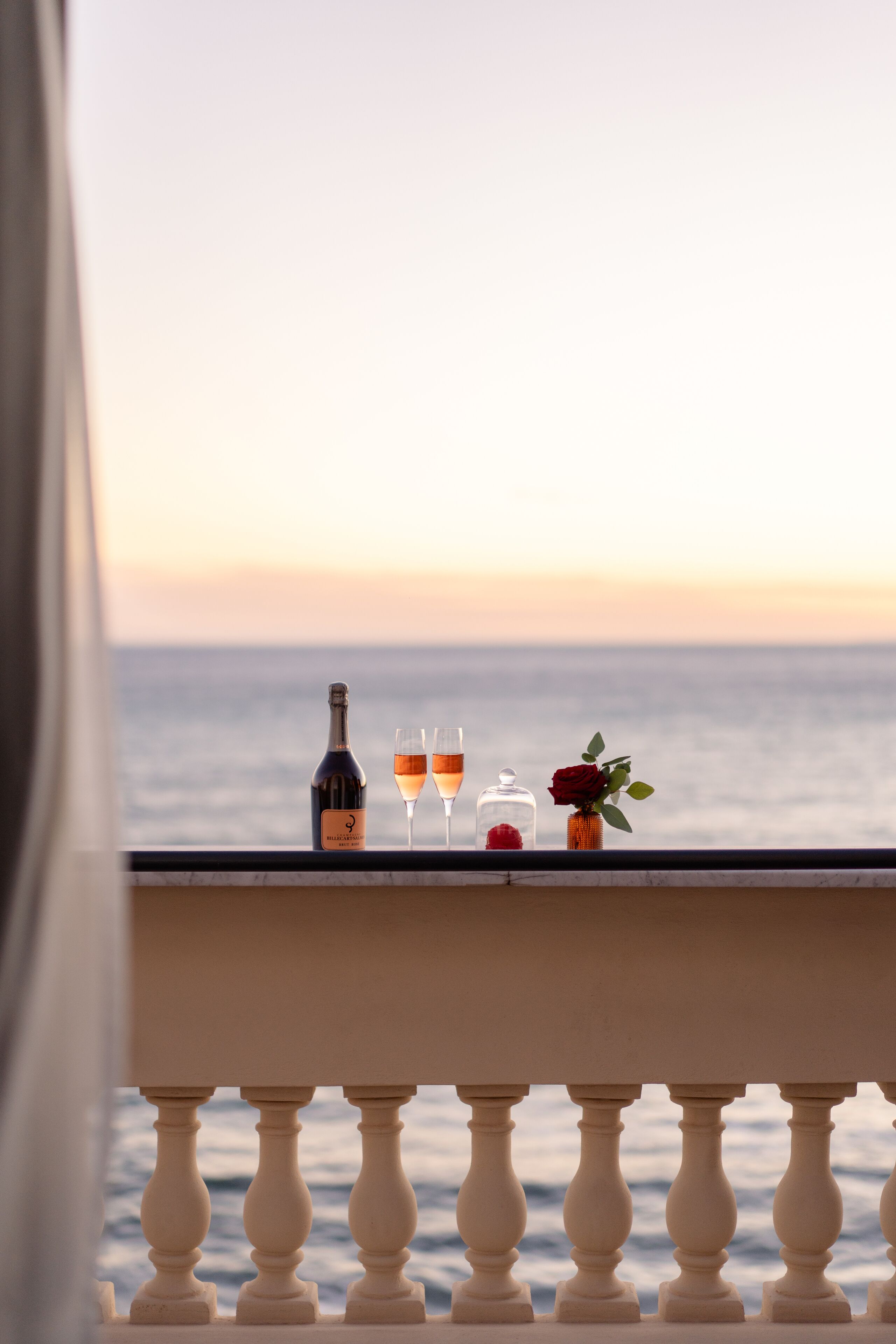 Un balcon avec vue sur la mer, une bouteille, deux verres et un vase avec une rose.